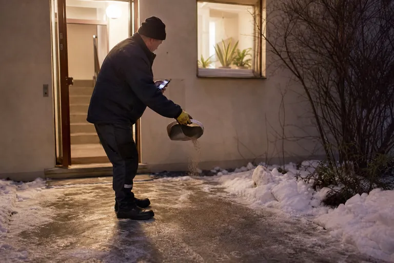 Hausmeister streut auf einem vereisten Gehweg vor einem Wohnhaus und dokumentiert den Einsatz am Smartphone.